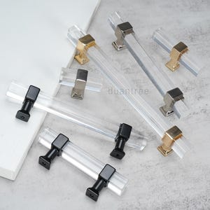 May include: A collection of cabinet pulls featuring clear, rectangular acrylic bars paired with metal mounting posts in black, silver, and gold finishes. The hardware is arranged in various lengths, showcasing a transparent design with solid metal bases.