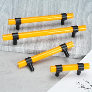 May include: Four cabinet pulls featuring bright yellow cylindrical bars and matte black mounting posts. Each handle has two black brackets securing the yellow bar, providing a consistent design across the varying lengths of the hardware pieces.