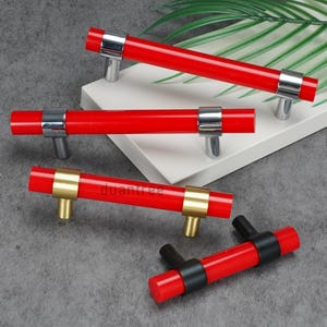 Könnte beinhalten: Rote Schrankgriffe mit silbernen, goldenen und schwarzen Akzenten. Die Griffe haben ein zylindrisches Design und sind auf einer weißen und grauen Oberfläche angeordnet. Das Wort "duantree" ist auf einem der Griffe aufgedruckt.