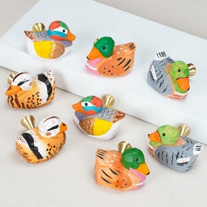 Peut inclure: Un ensemble de sept boutons de placard en bois en forme de canard peints à la main. Chaque bouton présente un motif de couleur unique, incluant des nuances d'orange, de vert, de jaune, de blanc et de gris, avec une tige de fixation dorée.