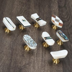 Può includere: Una collezione di pomelli decorativi per mobili con vari design. I pomelli presentano intarsi bianchi, iridescenti e a mosaico, montati su basi in ottone. Le forme variano, includendo design rettangolari, ovali e curvi, offrendo una gamma di opzioni estetiche per i mobili.