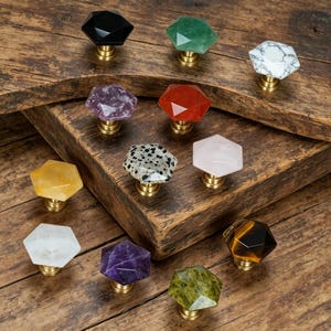 May include: A collection of gemstone cabinet knobs in various colors, including black, green, white, purple, red, yellow, and pink. Each knob has a faceted, hexagonal shape and a brass base. The knobs are displayed on a rustic wooden surface.