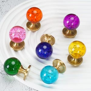 May include: A collection of seven spherical cabinet knobs featuring translucent glass balls with internal air bubbles, each mounted on a circular brushed brass base. The knobs are coloured in orange, purple, pink, dark blue, yellow, green, and light blue.