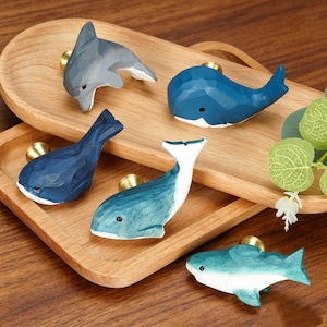 May include: Five drawer knobs shaped like marine animals, including a gray dolphin, a dark blue whale, a navy blue whale, a teal whale, and a light blue whale. Each knob features a gold-toned metal base for attachment to furniture surfaces.