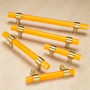 May include: A collection of five cabinet pulls featuring bright orange cylindrical bars accented with brushed gold metal mounting brackets. The hardware set includes various lengths of handles, each designed with two circular posts for attachment.