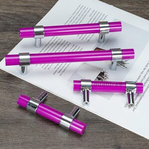 May include: Several magenta cabinet handles with a cylindrical design. Each handle has a silver-toned metal base and mounts. The handles are displayed on a white surface with printed text.