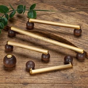 May include: A collection of brass cabinet pulls with dark brown wooden knobs. The pulls have a rectangular shape with rounded edges. The knobs are spherical. The hardware is arranged on a wooden surface with a sprig of greenery.