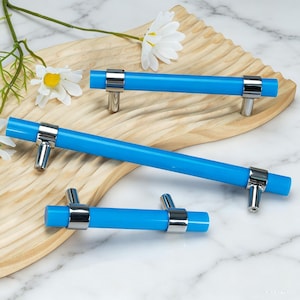 May include: Three bright blue cylindrical cabinet pulls with polished chrome metal mounting brackets. The hardware features a smooth, solid-coloured bar design with two silver-toned supports on each piece, varying in length across the set.