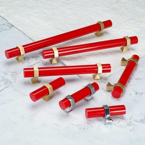 May include: A collection of cylindrical cabinet pulls in bright red with metallic accents. The pulls feature gold-toned or silver-toned hexagonal mounting brackets that secure the red rods. These hardware pieces vary in length and orientation.