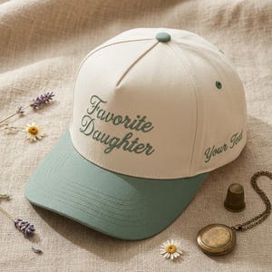 May include: A cream-colored baseball cap with a green brim and button. The words "Favorite Daughter" are embroidered in green script on the front. The text "Your Text" is embroidered on the side. A stylish accessory.