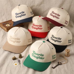May include: A collection of baseball caps in various colors, including navy, red, green, and black. Each cap features the embroidered text "Favorite Daughter" and "Your Text" on the side. The caps are displayed on a neutral fabric.
