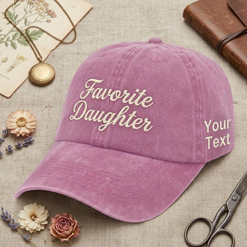 May include: A dusty rose baseball cap with the words "Favorite Daughter" embroidered in a cursive font. The cap also has the words "Your Text" on the side. The hat is a casual accessory.