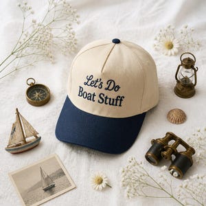 May include: A two-tone baseball cap with a cream-coloured crown and a navy blue brim. The front of the crown features the embroidered text Let's Do Boat Stuff in navy blue script and block lettering. The cap has a small navy blue button on top.