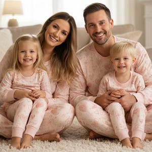 Könnte beinhalten: Eine vierköpfige Familie in passenden rosa Pyjamas mit Herzmuster sitzt auf einem weißen Teppich. Die Eltern und zwei Kinder lächeln in die Kamera. Der Hintergrund umfasst eine Couch, einen Kamin und eine weiße Wand.