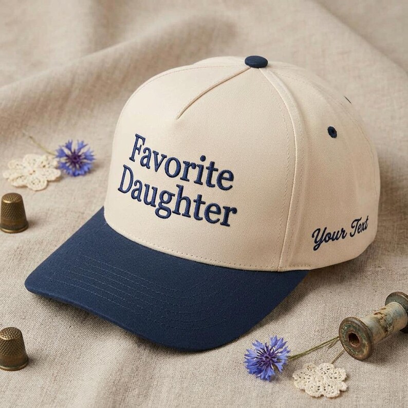 May include: A baseball cap with a cream-colored crown and a navy blue brim. The text "Favorite Daughter" is embroidered in navy blue on the front. The words "Your Text" are embroidered on the side.