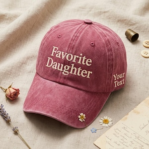 May include: A maroon baseball cap with the embroidered text "Favorite Daughter" in white. The cap also features the text "Your Text" and is decorated with small flowers. A thimble and buttons are in the background.