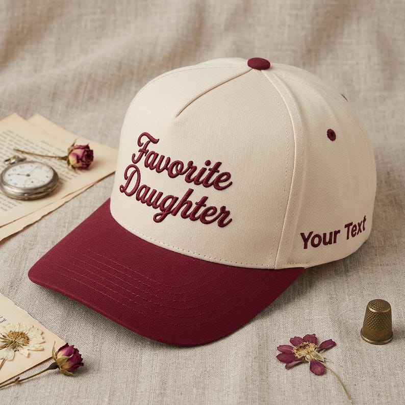 May include: Cream-colored baseball cap with a burgundy brim and top button. The cap features the embroidered text "Favorite Daughter" in burgundy script. The right side of the cap has the text "Your Text".
