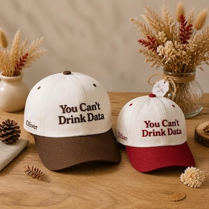 May include: Two baseball caps with white crowns and colored brims. The larger cap has a brown brim and brown embroidery reading "You Can't Drink Data" and "Oliver". The smaller cap has a maroon brim and maroon embroidery reading "You Can't Drink Data" and "Ethan".