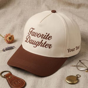 May include: A baseball cap with a brown brim and top button, and a cream-colored crown. The words "Favorite Daughter" are embroidered in brown script on the front. The text "Your Text" is embroidered on the side.