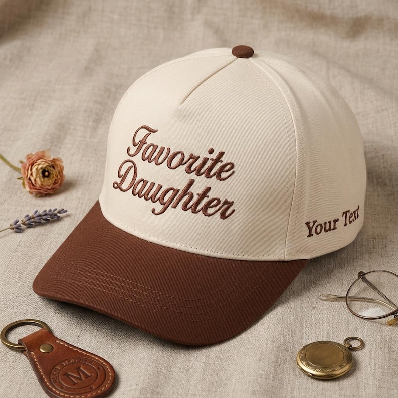 May include: A baseball cap with a brown brim and top button, and a cream-colored crown. The words "Favorite Daughter" are embroidered in brown script on the front. The text "Your Text" is embroidered on the side.