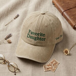 May include: A tan baseball cap with the embroidered text "Favorite Daughter" in green. The hat has a curved brim and a button on top. The words "Your Text" are embroidered on the side. The hat is on a tan surface with other items.