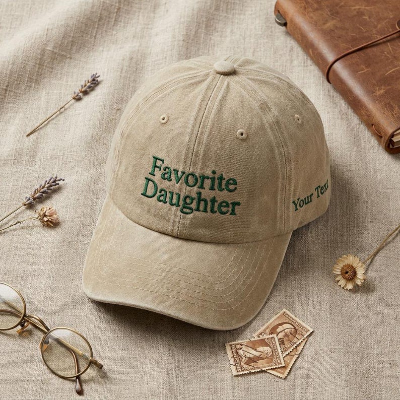 May include: A tan baseball cap with the embroidered text "Favorite Daughter" in green. The hat has a curved brim and a button on top. The words "Your Text" are embroidered on the side. The hat is on a tan surface with other items.