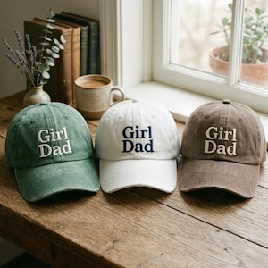 May include: Three baseball caps in green, white, and brown, each embroidered with "Girl Dad" in a contrasting color. The caps are displayed on a wooden surface, with a window and other items in the background.
