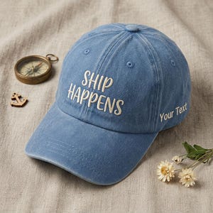 Könnte beinhalten: Blaue Baseballkappe mit dem gestickten Text "SHIP HAPPENS" in Weiß. Die Kappe hat auch den Text "Your Text" auf der Seite. Ein Kompass und ein Anker-förmiger Anhänger liegen neben der Kappe.