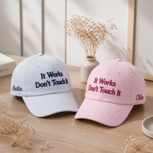 May include: A light blue and white pinstriped baseball cap with the dark blue embroidered text 'It Works Don't Touch It' and 'Austin' on the side, alongside a pink and white pinstriped baseball cap with dark pink text 'It Works Don't Touch It' and 'Chloe'.