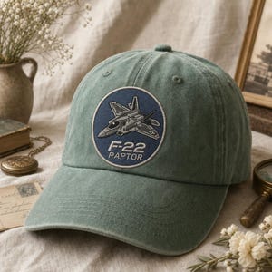 May include: A sage green baseball cap featuring a circular embroidered patch on the front. The patch displays a dark blue background with a detailed illustration of an F-22 Raptor fighter jet and the text F-22 RAPTOR in white lettering.