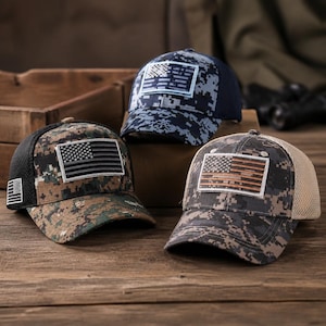 Pode incluir: Três bonés de beisebol com padrões de camuflagem digital e emblemas bordados da bandeira americana na frente. Um boné tem rede preta, um tem rede azul-marinho e um tem rede bege com um emblema de bandeira castanha.