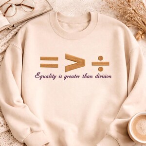 May include: A cream-coloured sweatshirt with the embroidered phrase "Equality is greater than division." The design features mathematical symbols: an equals sign, a greater-than sign, and a division symbol. The text is in a purple colour.
