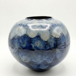 May include: A round ceramic vase with a dark interior and a textured exterior. The vase features a pattern of overlapping, scale-like shapes in shades of blue and white, creating a gradient effect. The vase has a glossy finish.