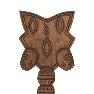 May include: A carved wooden artifact with a handle and a decorative top. The top features three oval shapes and two carved faces. The handle has a series of carved geometric patterns. The wood has a warm brown tone.