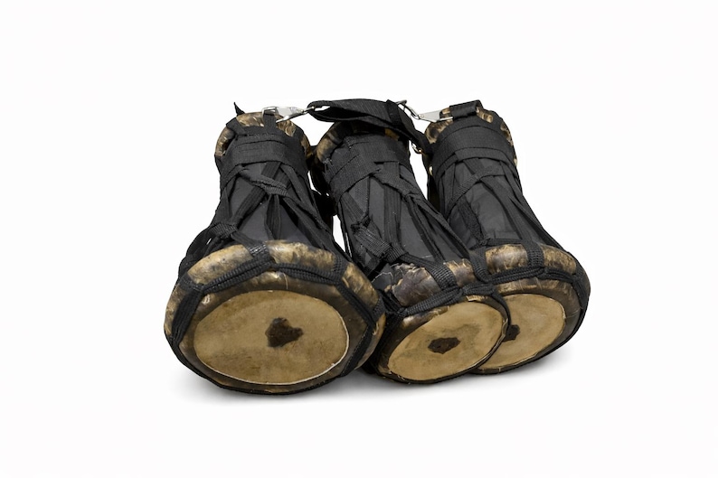 Peut inclure: Trois petits djemb&eacute;s avec du tissu noir tiss&eacute; et des peaux de tambour beiges. Les tambours sont reli&eacute;s par une attache m&eacute;tallique et des sangles noires. Les tambours sont pos&eacute;s sur un fond blanc.