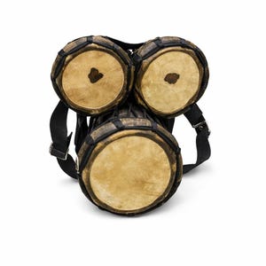 Peut inclure: Un ensemble de trois tambours bruns et noirs avec des peaux de tambour beiges. Les tambours sont reli&eacute;s et ont une sangle noire pour le transport. Les tambours sont en bois et en cuir.