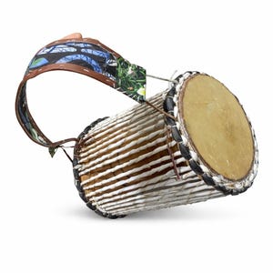 May include: A hand drum with a light brown drumhead and a white and brown woven body. The drum has a patterned strap with blue, green, and brown accents. The drum's rim is black and white.