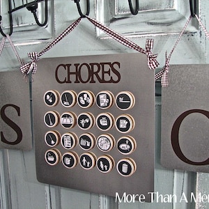 May include: A gray metal chore chart with the word "CHORES" at the top and the letters "S" and "C" on either side. The chart has circular icons representing different chores. The chart is suspended by brown and white checkered ribbon.