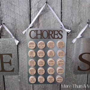 May include: A metal sign with the word "CHORES" in brown lettering.  The sign has a grid of 16 round, brown, metal tokens with text and dollar amounts.  The tokens are attached to the sign with magnets.  The sign is hanging from a hook on a wooden wall.  There are two smaller metal signs on either side of the chore sign, each with a letter, "E" and "S" in brown lettering.