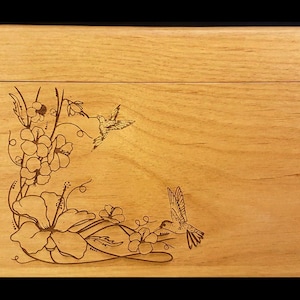 Könnte beinhalten: Eine Holzkiste mit einem Scharnierdeckel, verziert mit einem gravierten Design aus Hibiskusblüten und Kolibris. Die Kiste ist aus hellem Holz mit einer natürlichen Oberfläche.