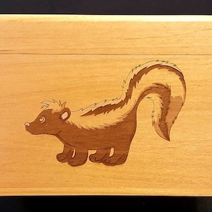 Könnte beinhalten: Eine Holzkiste mit einem Scharnierdeckel. Die Vorderseite der Kiste zeigt ein Cartoon-Bild eines Stinktiers mit einem schwarz-weiß gestreiften Schwanz.