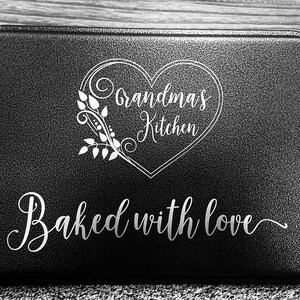 May include: Black kitchen mat with white text that reads "Grandma's Kitchen" inside a heart shape with a floral design. The text "Baked with love" is below the heart.