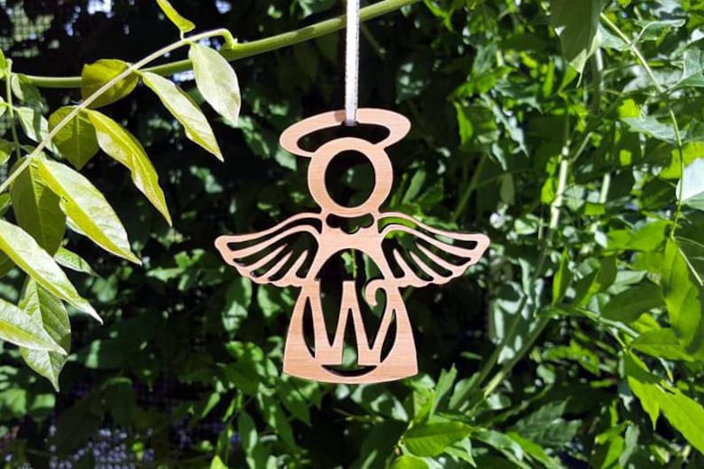 May include: Wooden angel ornament with a halo and wings, featuring a cutout letter "W" in the centre. The ornament is light brown and is hanging from a string.
