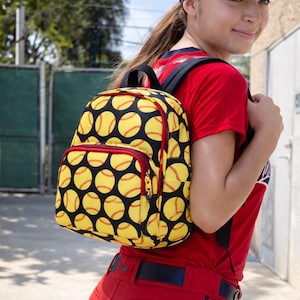 May include: A yellow and black softball-themed backpack with a red zipper and front pocket. The backpack is covered in a pattern of yellow softballs with red stitching. The person is wearing a red shirt and pants.