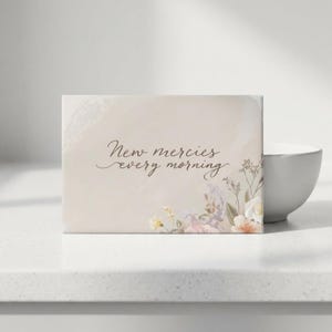 May include: A rectangular card with the text "New mercies every morning" in elegant script. The card features a soft, neutral background with floral accents in shades of pink, white, and yellow.
