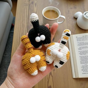 Peut inclure: Trois ornements faits à la main en crochet représentant des derrières de chats, en noir, orange et blanc, avec des pompons blancs duveteux. Les derrières de chats sont tenus dans une main, avec une tasse de café et un livre en arrière-plan.