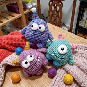 Könnte beinhalten: Drei handgefertigte Häkelmonster-Spielzeuge in Lila, Türkis und Rosa. Jedes Monster hat große weiße Augen, einen lächelnden Mund und eine Partyhütchen. Die Spielzeuge werden auf einer Holzoberfläche mit Wollknäueln und einer rosa Decke präsentiert.