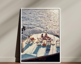 May include: A framed print showing a sunny seaside scene. Three people relax on deck chairs under striped umbrellas. The ocean glistens in the background, with a wooden floor and a neutral wall in the foreground.
