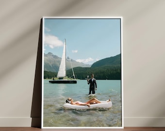 May include: A framed print showing a lake scene with a sailboat and mountains. A person floats on a white inflatable, while another in a suit wades in the water holding a tray with drinks, a tranquil scene.