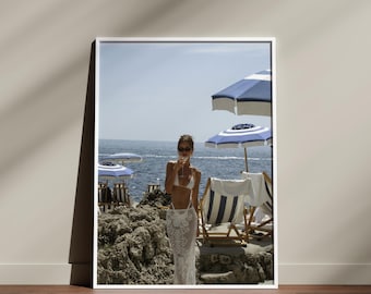 May include: A framed photograph depicts a beach scene with blue and white striped umbrellas, lounge chairs, and a person standing near the water. The person is wearing sunglasses and a white outfit. The ocean and sky are visible in the background.